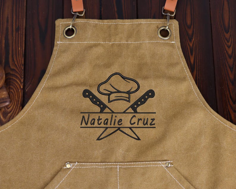 May include: Tan canvas apron with brown leather straps and metal hardware. The apron features a chef hat and crossed knives graphic with the name "Natalie Cruz" in black. A pocket is visible at the bottom.
