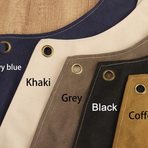 May include: Close-up of aprons in various colors: navy blue, khaki, grey, black, and coffee. Each apron has a metal grommet and is labeled with its color. The aprons are made of a sturdy fabric.
