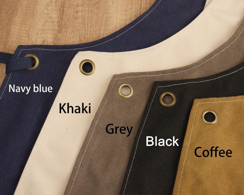 May include: Close-up of aprons in various colors: navy blue, khaki, grey, black, and coffee. Each apron has a metal grommet and is labeled with its color. The aprons are made of a sturdy fabric.