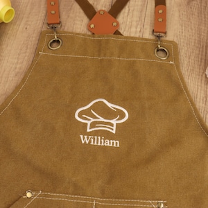 May include: Brown canvas apron with a white chef hat logo and the name "William" embroidered on the front. Features brown leather straps and metal hardware. A practical and personalized kitchen accessory.