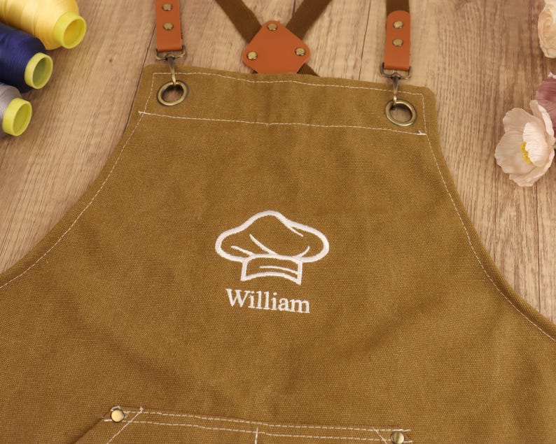 May include: Brown canvas apron with a white chef hat logo and the name "William" embroidered on the front. Features brown leather straps and metal hardware. A practical and personalized kitchen accessory.