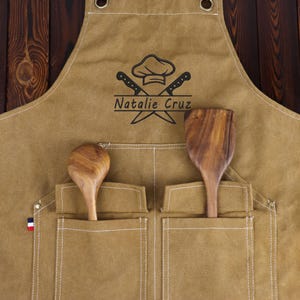 May include: A tan apron with two front pockets, holding a wooden spoon and spatula. The apron features a chef's hat and crossed knives logo, with the name "Natalie Cruz" below. Brown leather straps and a French flag detail are visible.