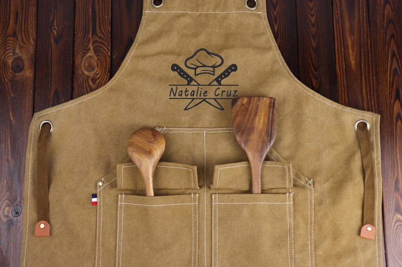 May include: A tan apron with two front pockets, holding a wooden spoon and spatula. The apron features a chef's hat and crossed knives logo, with the name "Natalie Cruz" below. Brown leather straps and a French flag detail are visible.