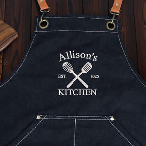 Pode incluir: Avental de cozinha em ganga azul escuro com costuras brancas e um bolso frontal. O avental apresenta alças de couro e ferragens metálicas. Bordado em branco estão as palavras "Allison's Kitchen" com um design de batedor e espátula e o ano de 2025.
