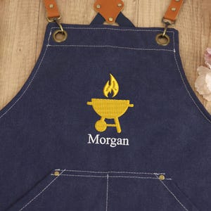 May include: A navy blue apron with brown leather straps and brass hardware. The apron features a yellow embroidered barbecue grill design with flames and the name "Morgan" in white. The apron has a front pocket and white stitching.