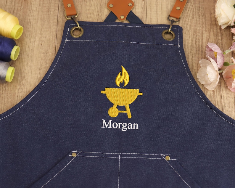 May include: A navy blue apron with brown leather straps and brass hardware. The apron features a yellow embroidered barbecue grill design with flames and the name "Morgan" in white. The apron has a front pocket and white stitching.