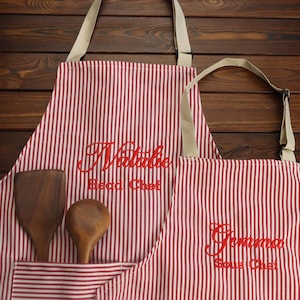 Puede incluir: Dos delantales a rayas rojas y blancas con los nombres "Natalie, Head Chef" y "Gemma, Sous Chef" bordados en rojo. Utensilios de cocina de madera están en el bolsillo del delantal. Los delantales tienen correas beige.