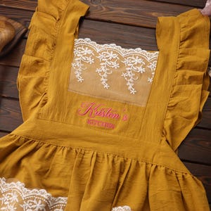 May include: Mustard yellow apron with white lace detailing and ruffled edges. The apron features the embroidered text "Kristen's Kitchen" in pink. Wooden spoons are visible in the background. A floral accent is on the right.