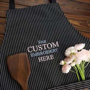 May include: Black and white striped apron with the text "Your Custom Embroidery Here" embroidered in blue and pink. A wooden spatula and a bouquet of pink flowers are in the apron pocket. The apron has black straps.