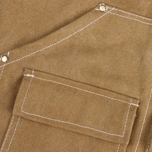 May include: Close-up of a tan canvas apron with white stitching and multiple pockets. The apron has a small patch with red, white, and blue stripes. Metal rivets reinforce the corners.