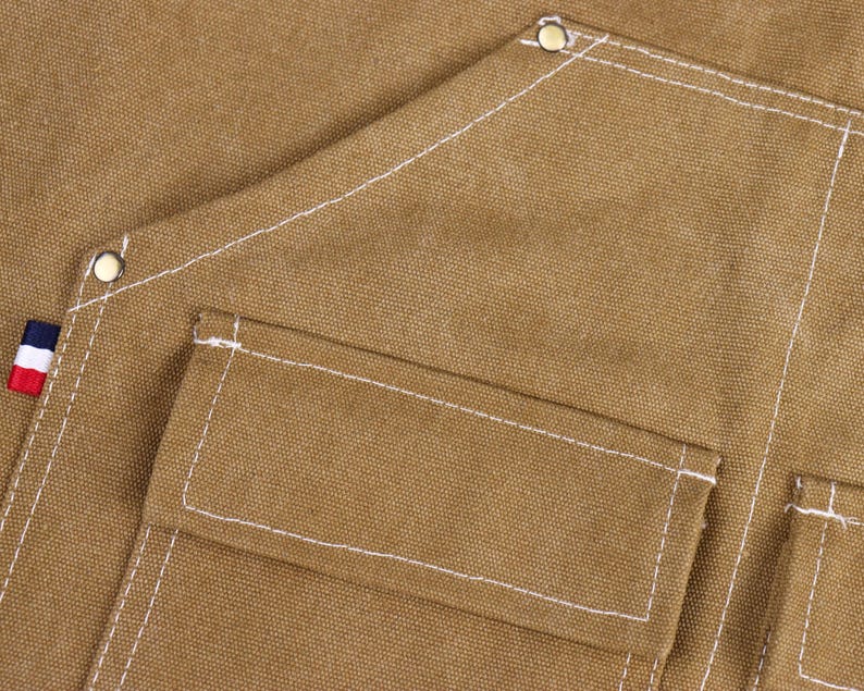 May include: Close-up of a tan canvas apron with white stitching and multiple pockets. The apron has a small patch with red, white, and blue stripes. Metal rivets reinforce the corners.