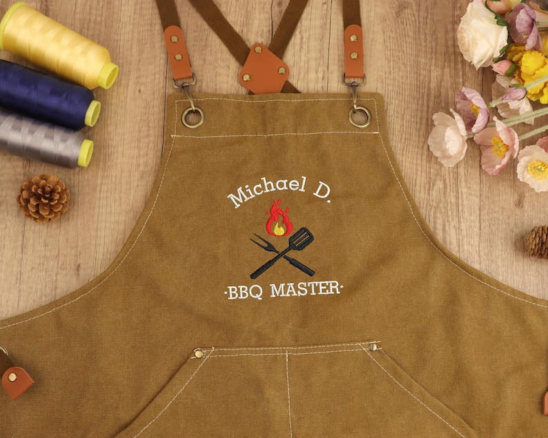 May include: A brown apron with leather straps and the embroidered text "Michael D. BBQ MASTER" above a crossed fork and spatula design with flames. The apron has a front pocket and metal hardware.