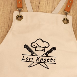 May include: Cream-colored apron with brown leather straps and metal hardware. The apron features a chef hat and crossed knives graphic with the name "Lori Knotts" in black. The apron is designed for kitchen use.