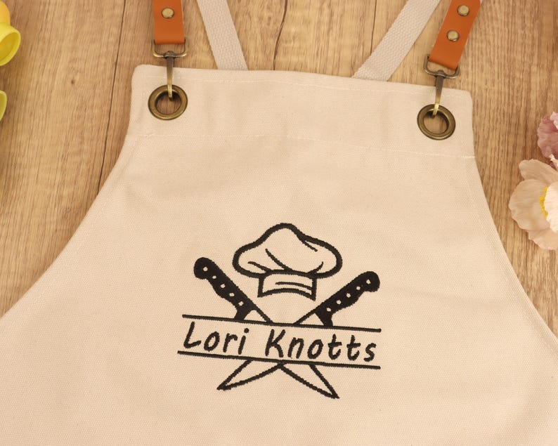 May include: Cream-colored apron with brown leather straps and metal hardware. The apron features a chef hat and crossed knives graphic with the name "Lori Knotts" in black. The apron is designed for kitchen use.
