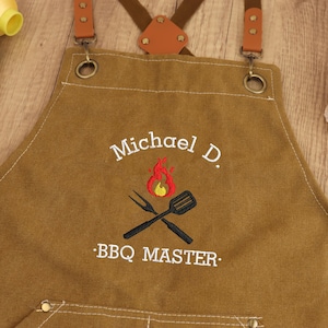 May include: Brown canvas apron with leather straps and metal hardware. The apron features embroidered text that reads "Michael D." above a flame and crossed grill tools, with "BBQ MASTER" below. The apron is designed for grilling.