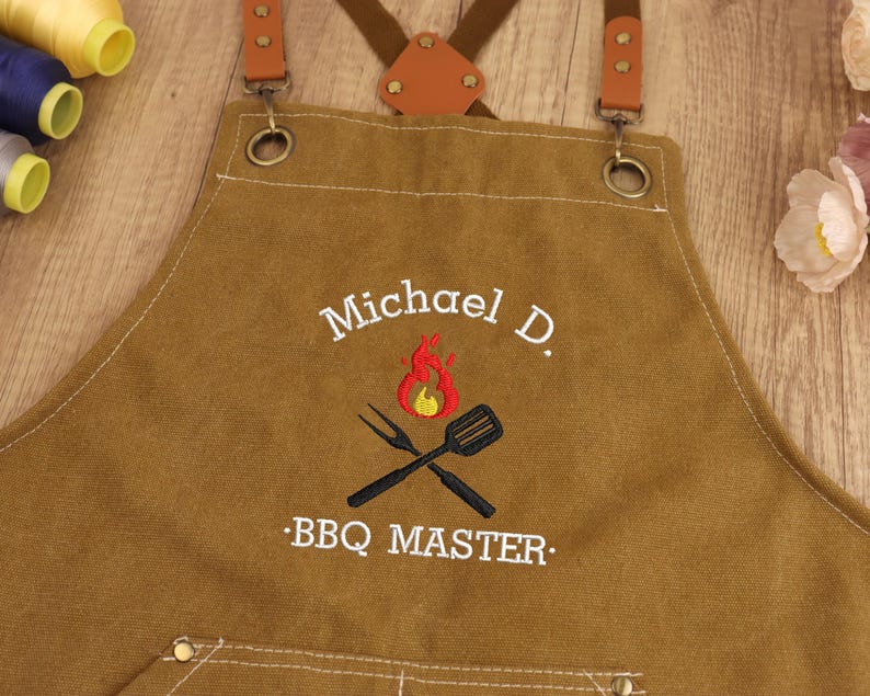May include: Brown canvas apron with leather straps and metal hardware. The apron features embroidered text that reads "Michael D." above a flame and crossed grill tools, with "BBQ MASTER" below. The apron is designed for grilling.