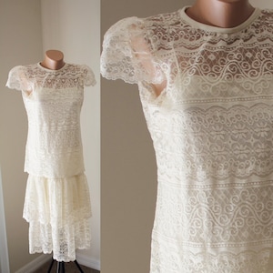 May include: A white lace dress with a tiered skirt and short sleeves. The dress has a delicate floral pattern and a round neckline.