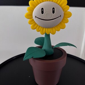Op de afbeelding: Een 3D-geprinte zonnebloem met een glimlachend gezicht, gele bloemblaadjes en groene bladeren, in een bruine pot.