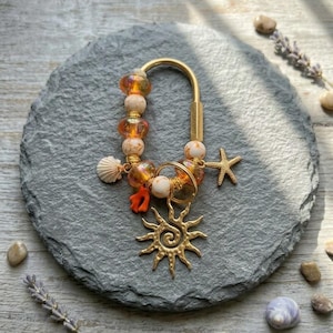 May include: A gold-toned metal keychain featuring a U-shaped clasp adorned with orange glass beads, white marbled beads, a small seashell charm, a bright orange coral-shaped charm, a gold starfish charm, and a large gold sun-shaped pendant.