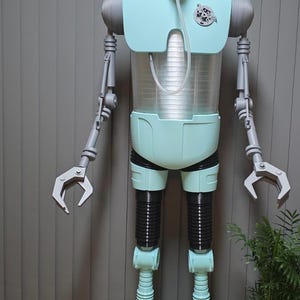 Può includere: Una figura robot a grandezza naturale con testa, busto e piedi azzurri, braccia articolate grigie e sezioni delle gambe nere a coste. Il robot presenta una sezione centrale trasparente, una maschera respiratoria sul viso e un emblema circolare.
