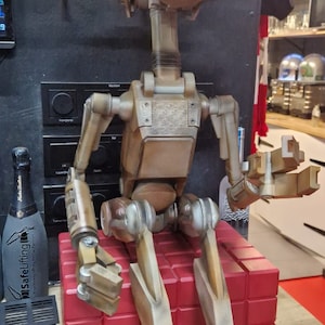 May include: A brown and beige robot with a large, round head and a white face. The robot is sitting on a red block with a yellow neon sign that says "STAR WARS" in the background.