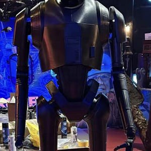 Puede incluir: Un robot negro y plateado con ojos azules. El robot tiene un cuerpo humanoide con articulaciones. El robot está de pie sobre una superficie roja.