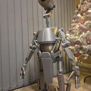 May include: A detailed metallic robot sculpture with a cylindrical head and articulated limbs. The robot features silver and gold accents, with a large, dark opening in its head. It stands on a wooden floor, with a Christmas tree visible in the background.