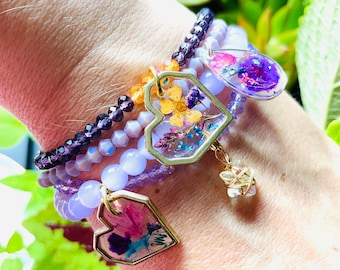 May include: A stack of purple, lavender, and clear beaded bracelets with gold heart-shaped charms. Each charm features pressed flowers and leaves encased in resin.