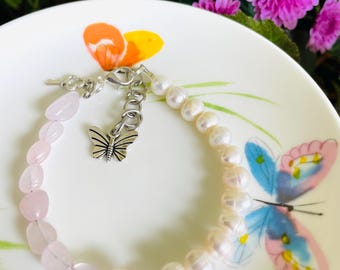May include: A delicate bracelet featuring a combination of pink rose quartz beads and white pearls. A silver butterfly charm hangs from the clasp. The bracelet is displayed on a white plate with a butterfly design, surrounded by pink flowers.