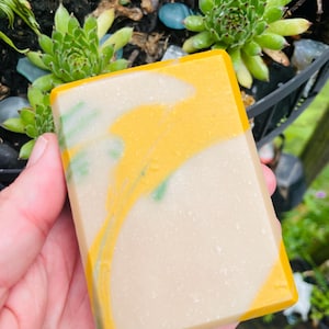May include: A rectangular bar of soap featuring a marbled design with swirls of bright yellow and a soft beige base color. The surface of the soap is smooth and solid, displaying a distinct color transition between the yellow and beige sections.