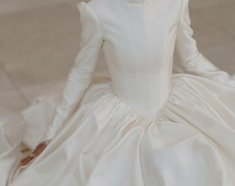 May include: A white wedding dress with a high neckline and long sleeves. The dress features a fitted bodice and a full skirt. The model is wearing a white head covering. The dress is made of a smooth, possibly satin, fabric.