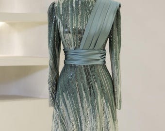 May include: A long-sleeved, sequined gown in shades of green and silver. The gown features a high neckline, a fitted bodice, and a flowing skirt. A matching sash is tied around the waist. The gown has a draped detail on one shoulder.