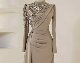 May include: A taupe-colored formal gown with long sleeves and a high neckline. The dress features intricate beading around the neckline and on one sleeve. The bodice has draped fabric details, and the skirt flows to the floor.