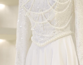 May include: A white wedding dress with long sleeves and a high neckline. The bodice is embellished with sequins and a pearl design. The skirt is a flowing, solid white fabric. The dress is elegant and suitable for a special occasion.