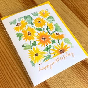 Puede incluir: Una tarjeta blanca con un borde amarillo y una ilustraci&oacute;n de acuarela de girasoles amarillos con hojas verdes. El texto "happy mother's day" est&aacute; escrito en cursiva naranja.