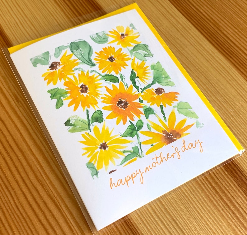 Puede incluir: Una tarjeta blanca con un borde amarillo y una ilustraci&oacute;n de acuarela de girasoles amarillos con hojas verdes. El texto "happy mother's day" est&aacute; escrito en cursiva naranja.