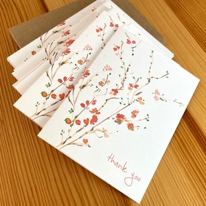 May include: A stack of thank you cards with a watercolor illustration of a branch with red and pink flowers. The text "thank you" is written in cursive on the front of the card.
