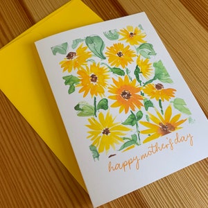 Puede incluir: Una tarjeta blanca con una pintura de acuarela de girasoles amarillos y hojas verdes. El texto "happy mother's day" est&aacute; escrito en cursiva en la parte inferior de la tarjeta.