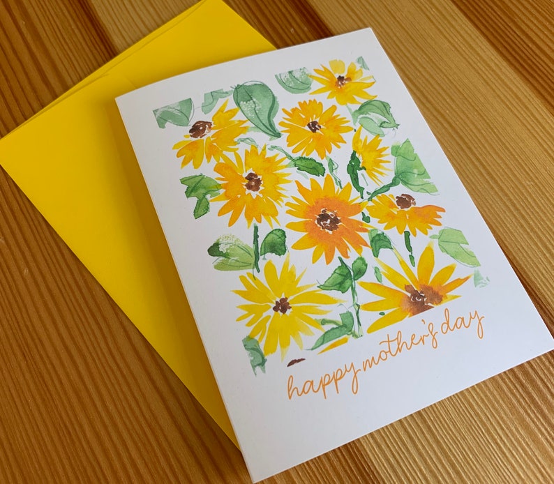 Puede incluir: Una tarjeta blanca con una pintura de acuarela de girasoles amarillos y hojas verdes. El texto "happy mother's day" est&aacute; escrito en cursiva en la parte inferior de la tarjeta.