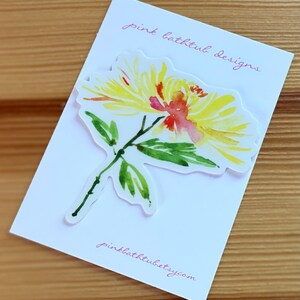 以下が含まれることがあります： ピンクの中心を持つ黄色の花の水彩画。花は、「ピンクバスタブデザイン」と「pinkbathtubetsy.com」というピンクのテキストが書かれた白いカードの上にあります。