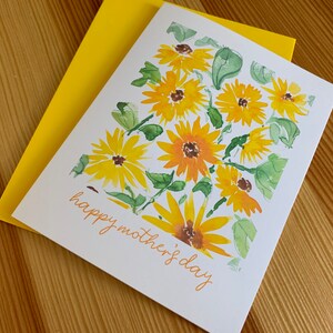 Puede incluir: Una tarjeta blanca con un sobre amarillo. La tarjeta tiene una pintura de acuarela de girasoles amarillos con hojas verdes y el texto "happy mother's day" en naranja.
