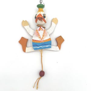 May include: A vintage wooden pull-string toy in the shape of a whimsical character. The toy features a white body, orange feet, and arms, with a painted face and a green hat. A red bead hangs from a string.