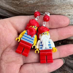 May include: Novelty earrings with Lego pirate figures. Each earring has a red hat, red trousers, and a yellow shirt with blue stripes. The earrings are connected with silver wire and red beads.