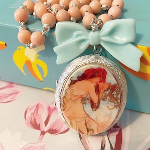 Può includere: Una collana con un ciondolo a forma di medaglione argentato, decorato con un fiocco blu pastello e una catena di perle rosa. Il medaglione presenta un'illustrazione vintage di una donna con i capelli rossi.