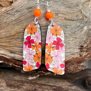 Può includere: Un paio di orecchini pendenti con perline arancioni e ciondoli floreali in legno. I ciondoli presentano uno sfondo bianco con fiori di ibisco rosa e arancioni.