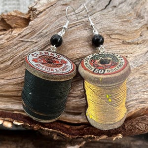 Può includere: Un paio di orecchini con bobine di filo e perline nere. Le bobine sono in legno e hanno etichette vintage. Una bobina è nera e l'altra è gialla.