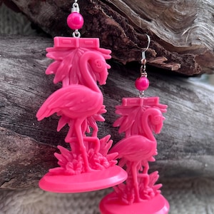Può includere: Un paio di orecchini a forma di fenicottero rosa acceso. Ogni orecchino presenta un fenicottero su una base circolare, con un motivo a palma dietro. Una perla rosa pende dall'amo d'argento. Gli orecchini sono in materiale liscio.