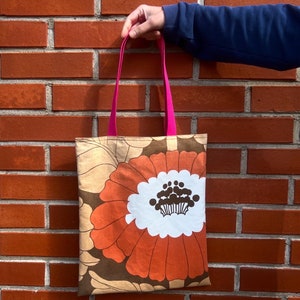 Puede incluir: Una bolsa de tela marrón y beige con un gran diseño de flor naranja. La bolsa tiene asas rosas.
