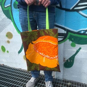 May include: A brown tote bag with green straps and a large orange flower with brown and green leaves printed on the front.
