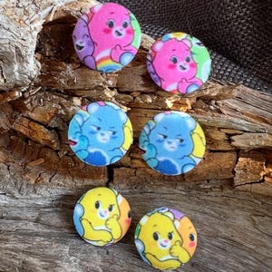 Può includere: Sei bottoni rotondi e colorati a tema Orsetti del Cuore. I bottoni presentano orsetti rosa, blu e gialli con sfondi arcobaleno, verdi e gialli. I bottoni sono disposti su un pezzo di legno stagionato.
